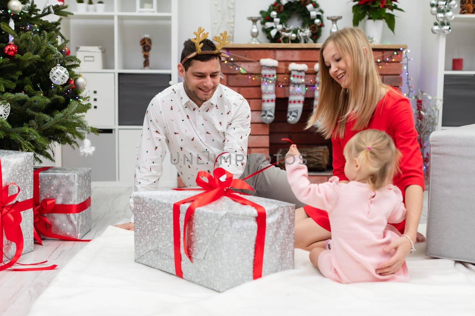 A woman, a man and a little girl sit in a room full of Christmas decorations. The whole family is sitting around a large gift. The box is wrapped with a red ribbon. The little girl pulls at the ribbon.