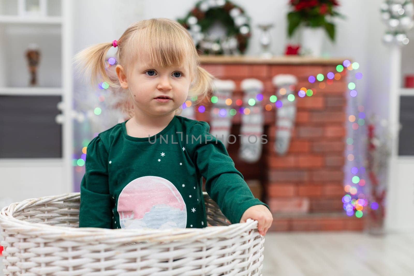 Close-up of a silhouette of a little girl sitting in a wicker basket. Blurred background. A brick fireplace can be seen in the background. Colorful Christmas lights are seen on the fireplace. The child is playing in the room.