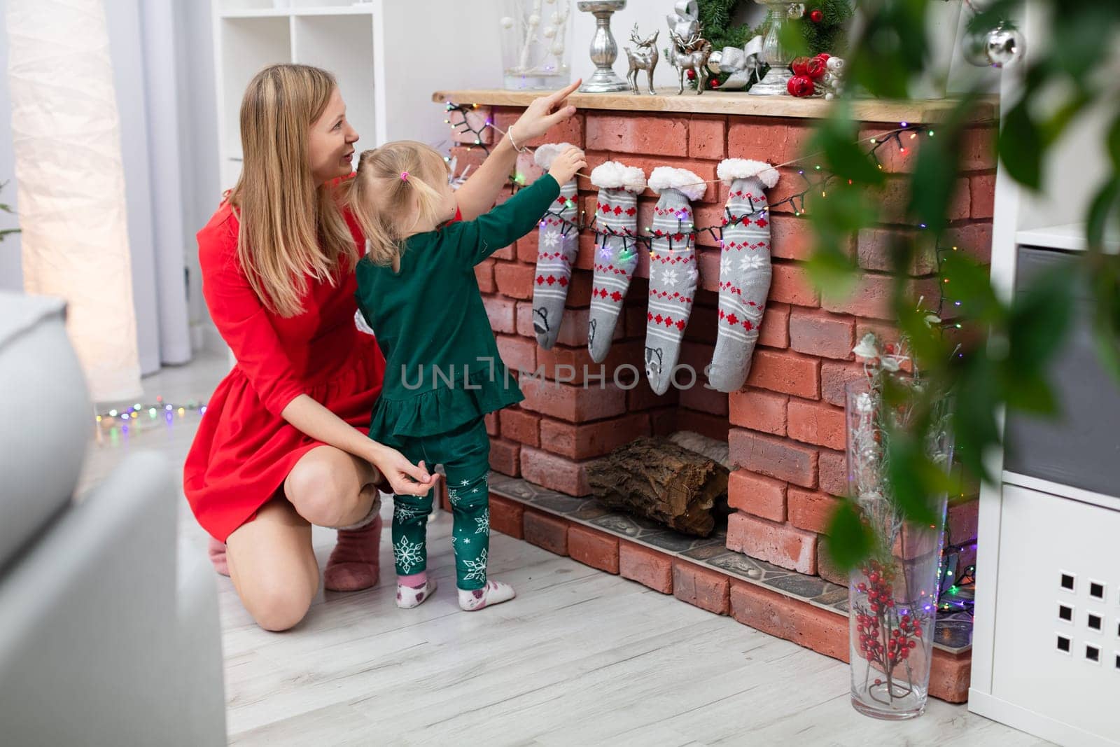 A woman dressed in a red dress crouches by the fireplace and embraces her daughter. The little girl stands by the fireplace and looks at the Christmas decorations. The brick fireplace is decorated with Christmas ornaments.