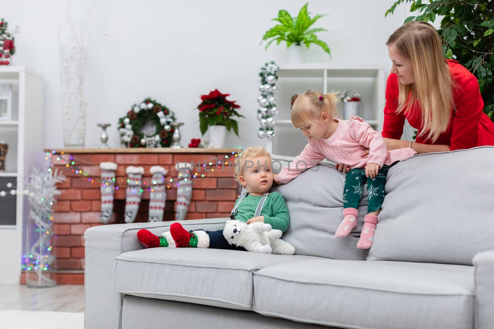 A little boy sits on a gray couch. A girl stands behind the boy and extends her hand toward him. Behind the couch stands a woman in a red dress. The mother is supporting her daughter to keep her from falling.