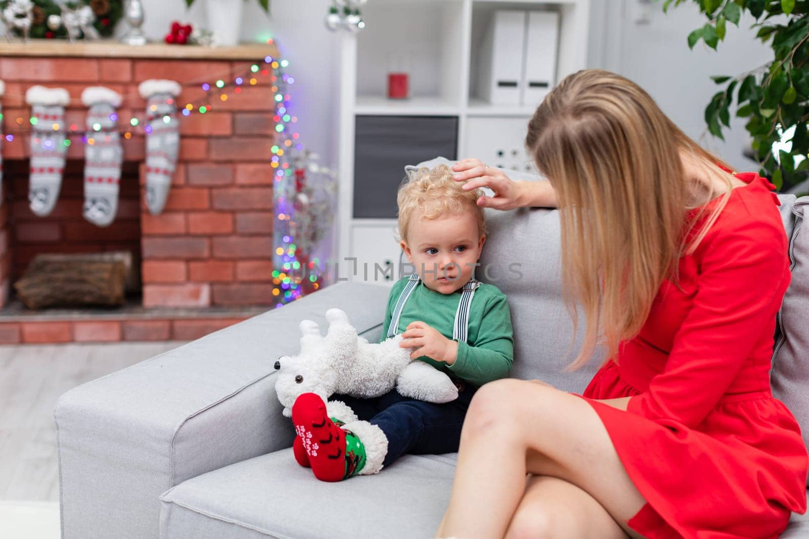 A young blonde woman sits on the couch with her little son. The mother is stroking her child's hair. The boy is holding a white cuddly toy. The room is decorated with Christmas decorations.