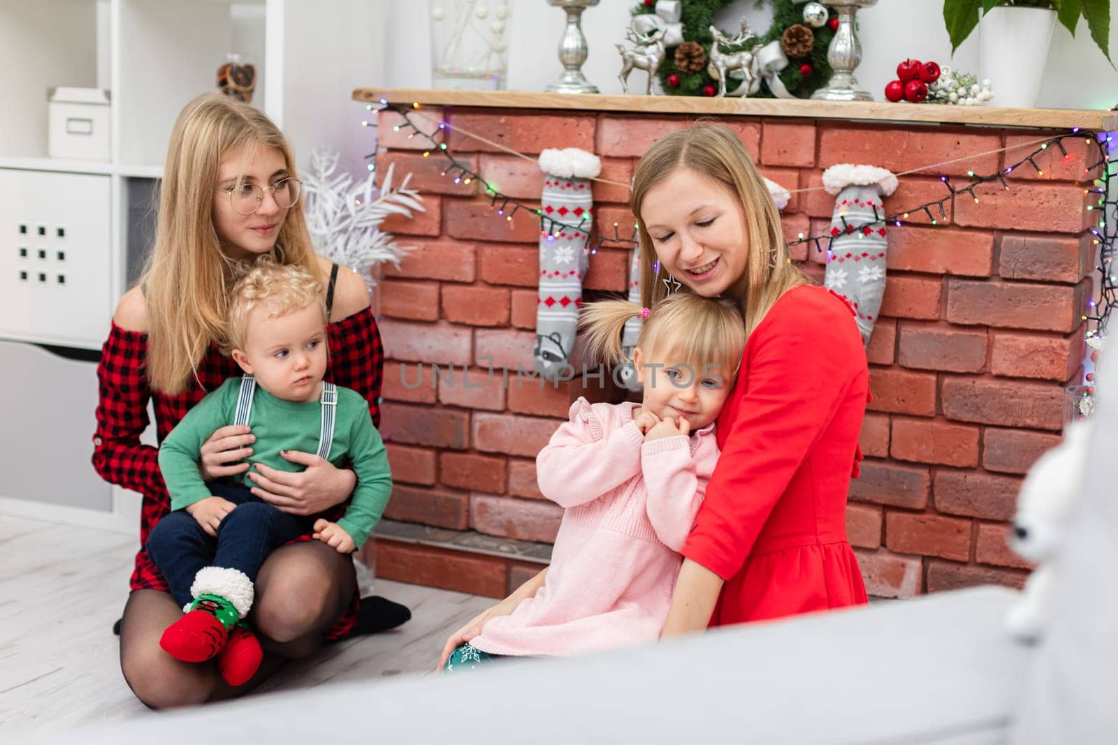 A woman in a red dress holds a little girl on her lap. The little girl is hugging her mother. Next to her sits a woman in glasses, who holds a little boy on her lap. The family is spending time by the fireplace.