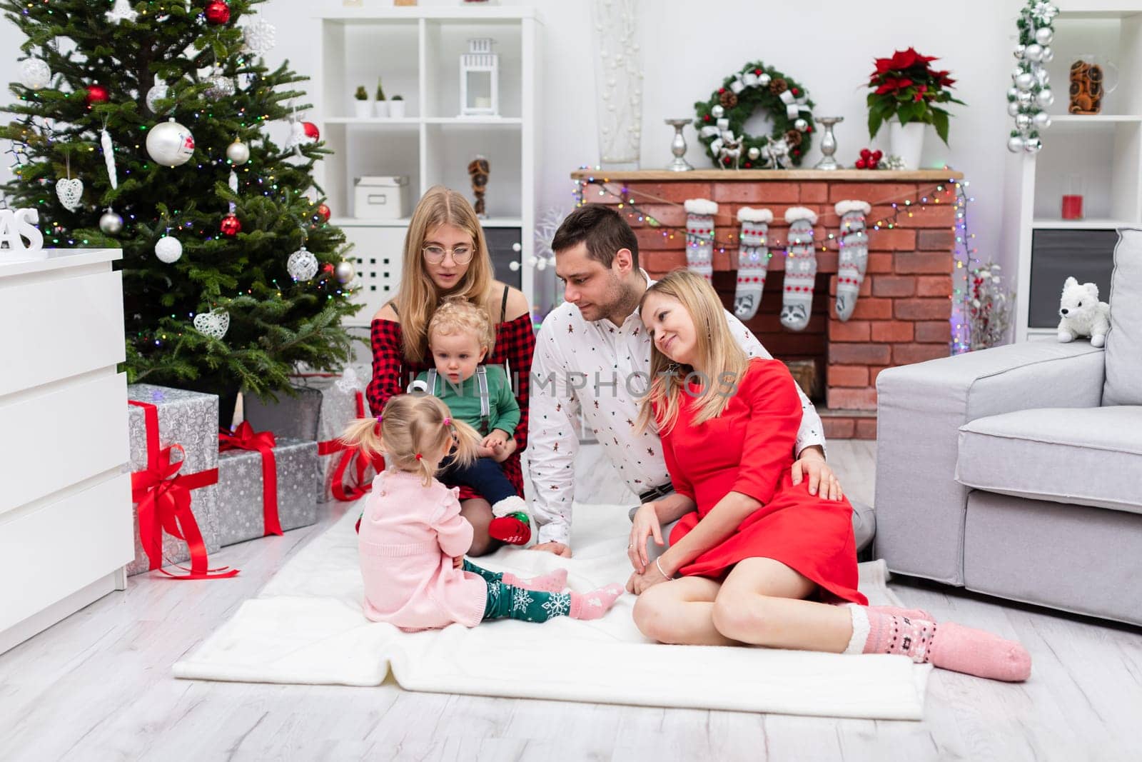 Two women, a man and two children spend Christmas together. The family sits on a white carpet in front of the fireplace. The fireplace is decorated with colorful lights. Presents lie under the large Christmas tree.