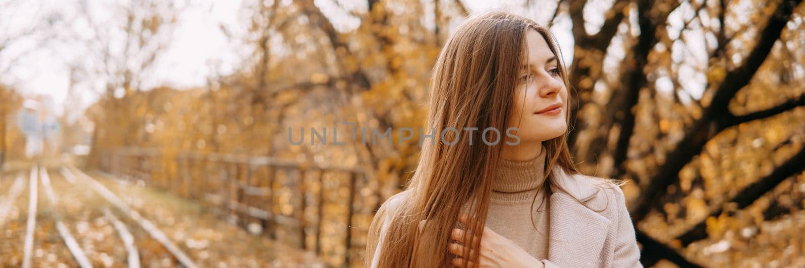 A beautiful long-haired woman walks through the autumn streets. Railway, autumn leaves, a woman in a light coat.