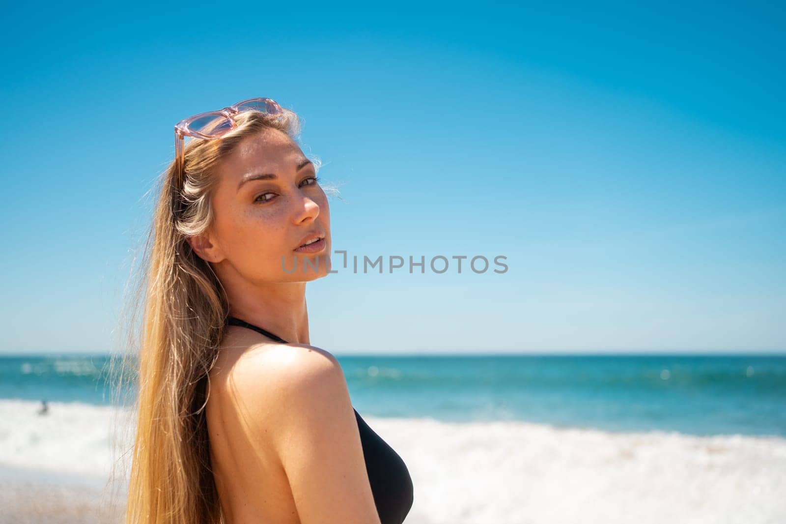 Blond woman on beach. Female with long hair and sunglasses on her head looking to camera while standing on sandy beach. Portrait of attractive girl in black swimwear with azure water and blue sky on backgrounds.