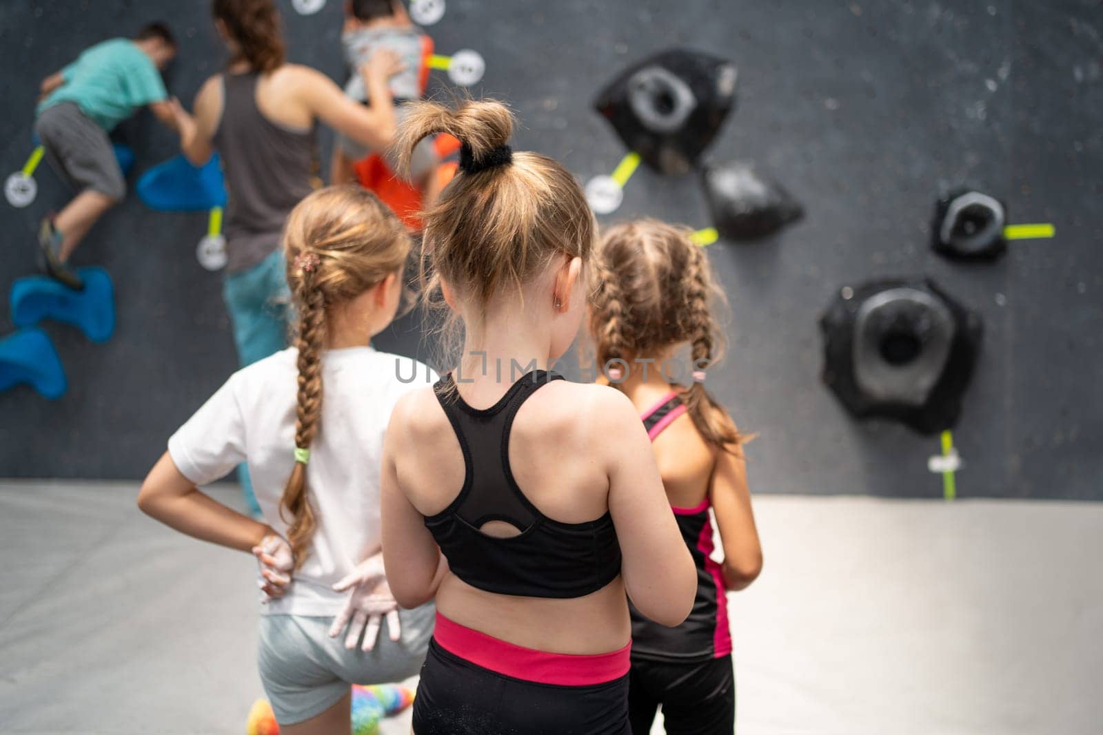 Three girls in sport clothes on climbing session. Back view of kids watching a coach in rock climbing class indoors. Young children looking on artificial boulder wall in gym