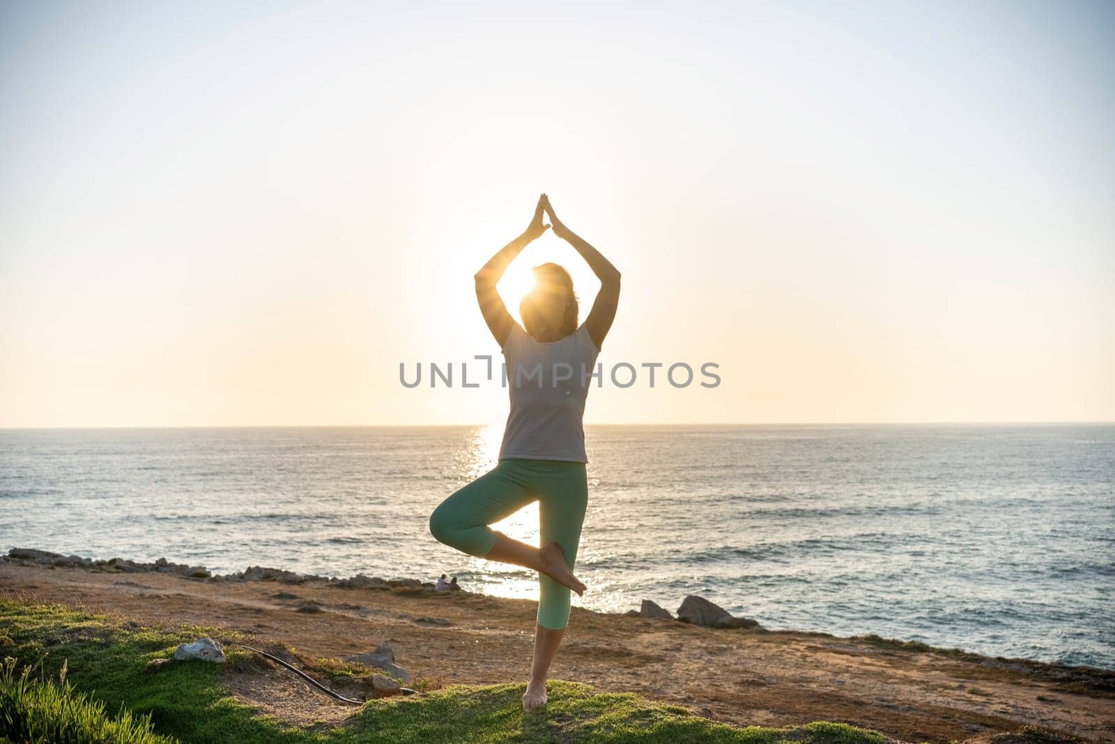 Senior woman practicing yoga on beach at sunset. Female doing yoga asana on coast with a sun setting on background. Silhouette of woman with slim body standing in yoga pose near ocean