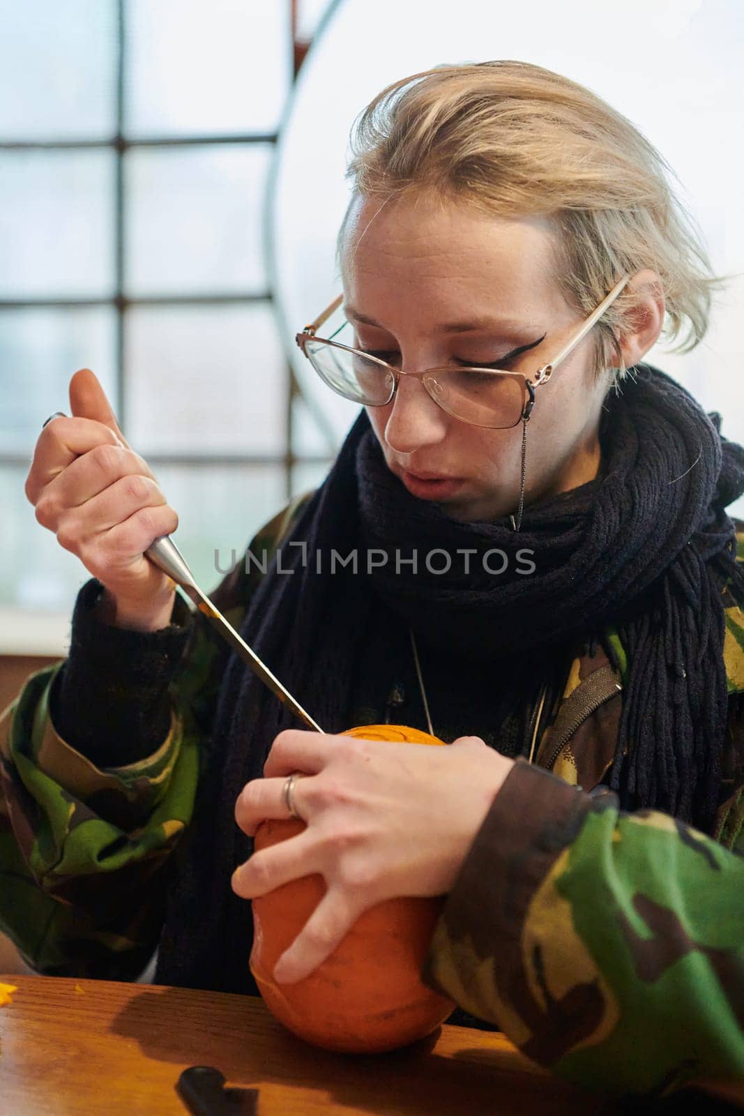 A modern blonde woman in military uniform is carving spooky pumpkins with a knife for Halloween night.