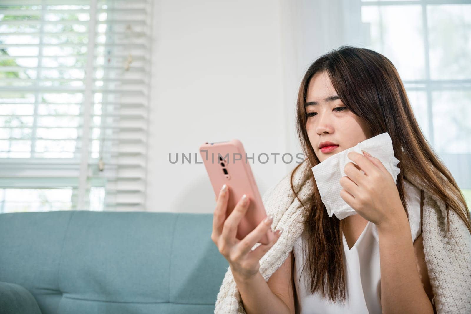 Asian young woman sick sitting down on sofa with cold blowing her nose and checking smart mobile phone to cell to doctor online, depressed sad sick female holding tissues and smartphone in living room