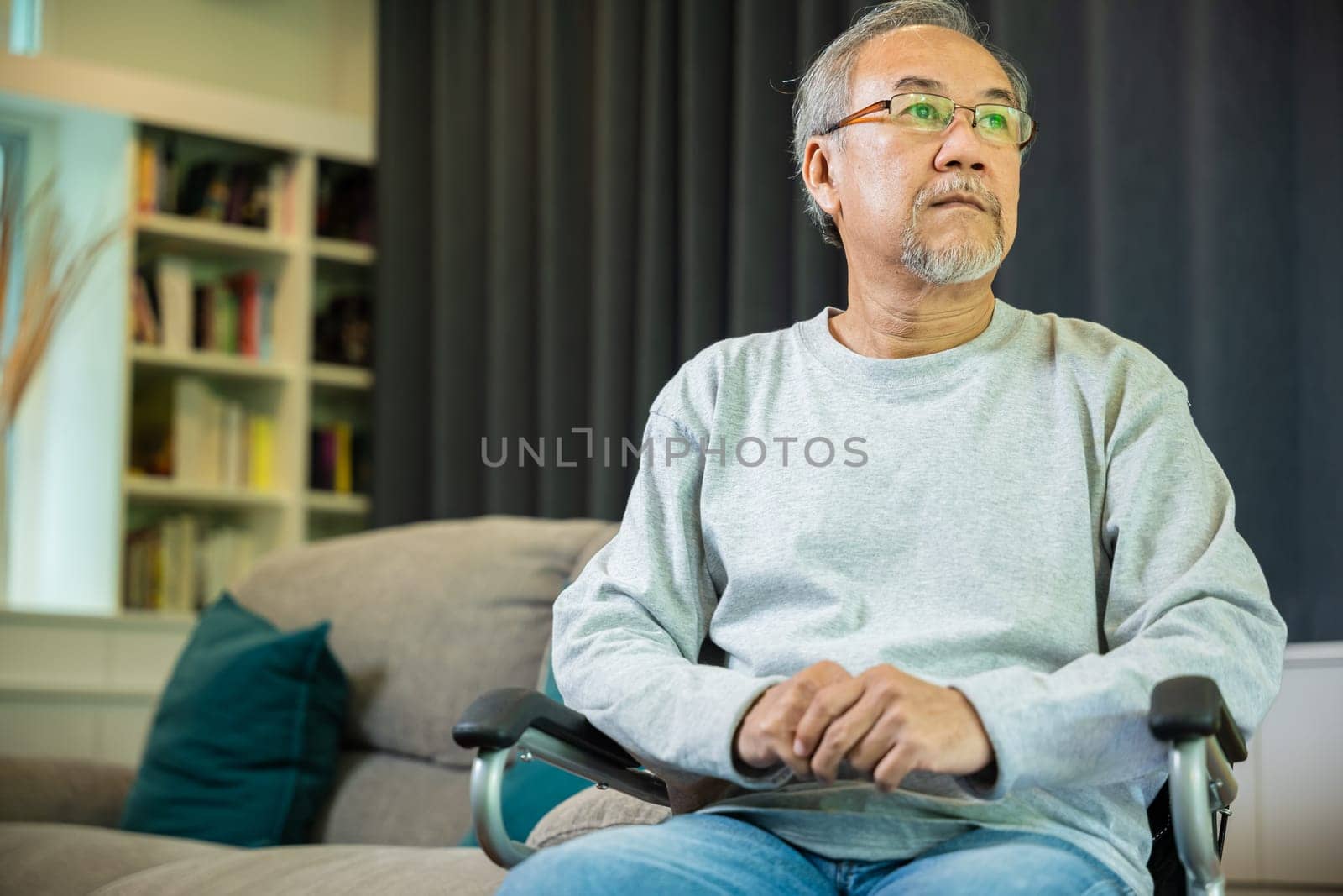 Asian senior man disabled sitting alone on wheelchair in living room looking out window at home and thinking, lonely elder sad old man retirement memory loss or nostalgia thought, feel depressed