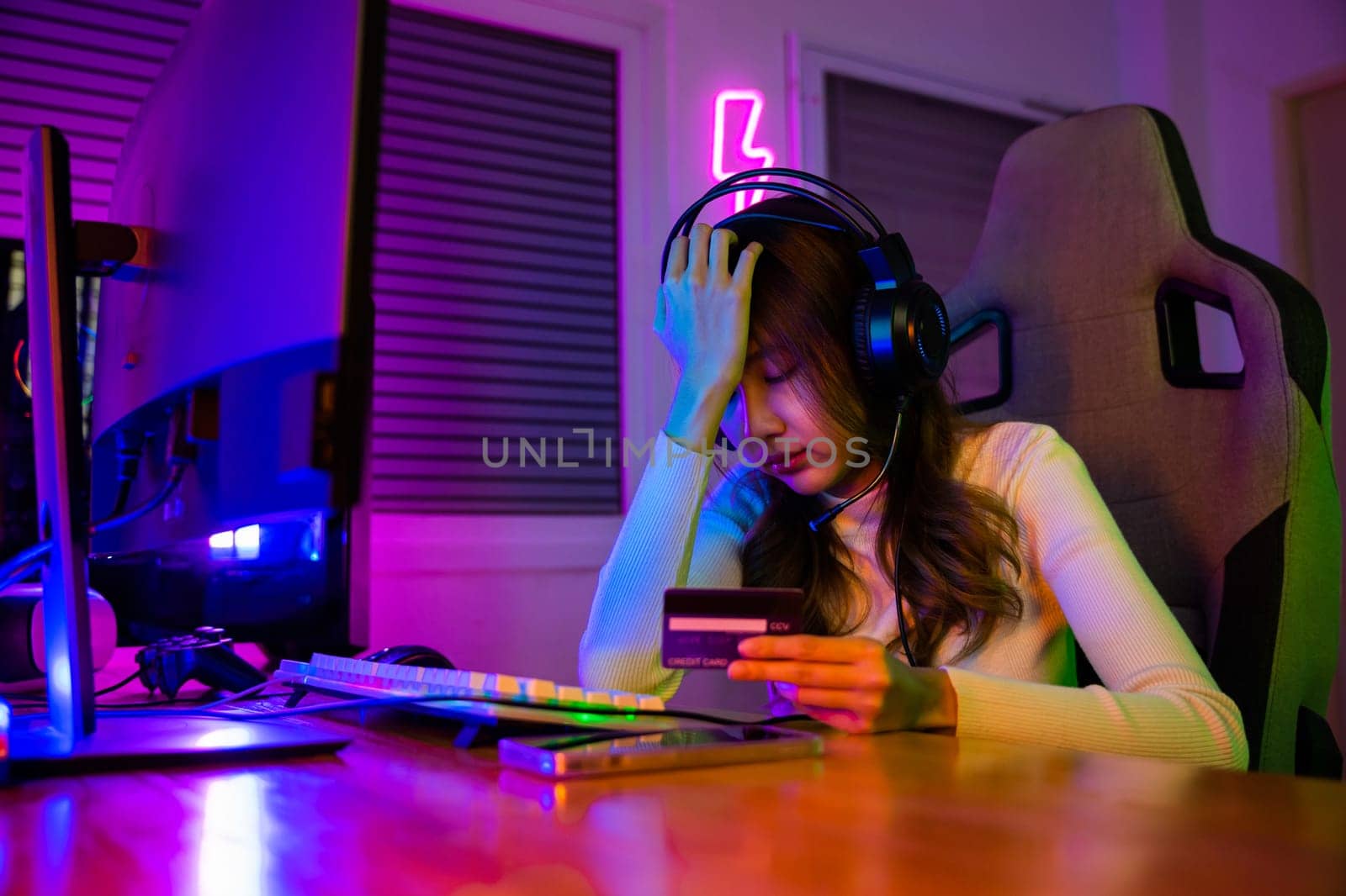 Young Asian woman gamer wearing headphones unset suffer of overspending money from card account after pay game online, problem using credit card, playing gaming with money