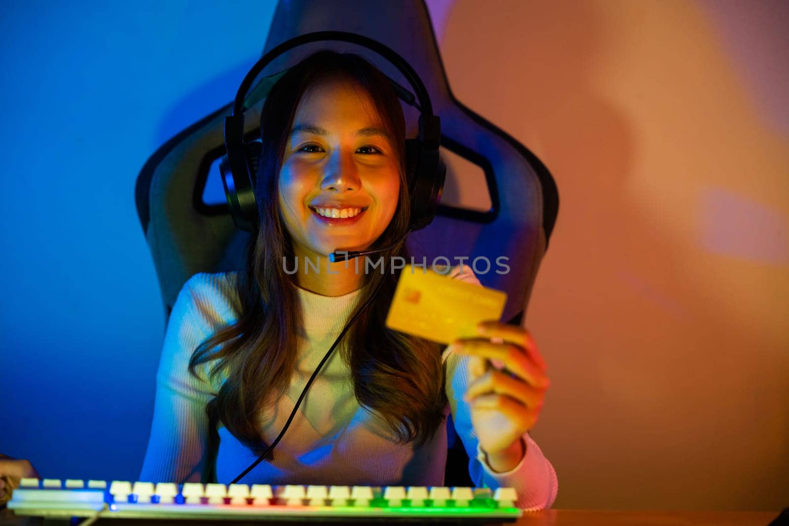 Asian gamer playing online video game and holding a credit card for payment game online, young woman in gaming headphones using computer for playing games at home, broadcast streaming live, Front view