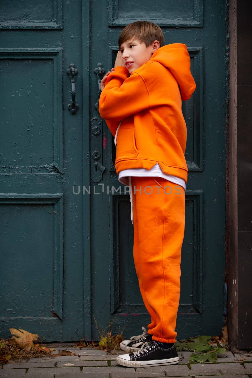 A teenage boy in an orange suit stands at the green door and eavesdrops.