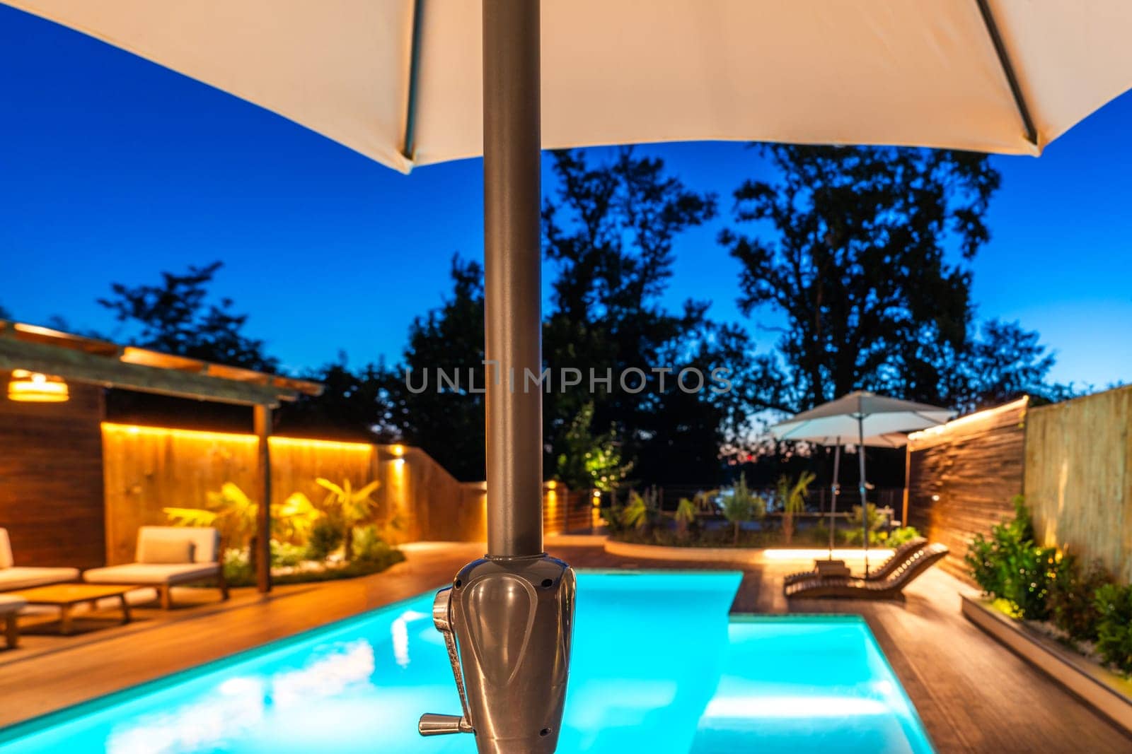 View of a luxurious backyard pool through a large umbrella under a twilight sky. Modern outdoor area with cozy lighting and sleek wooden decking