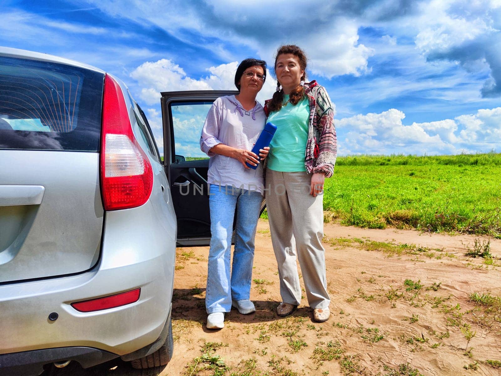 Women stand beside a silver car, enjoying the warm sun and expansive sky filled with clouds in a lush green field.