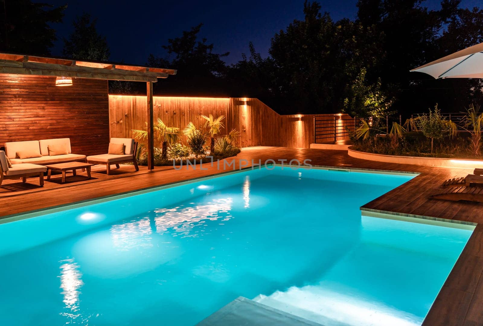 Modern pool with soft lighting, wooden deck, and loungers at night. Concept of peaceful outdoor escape