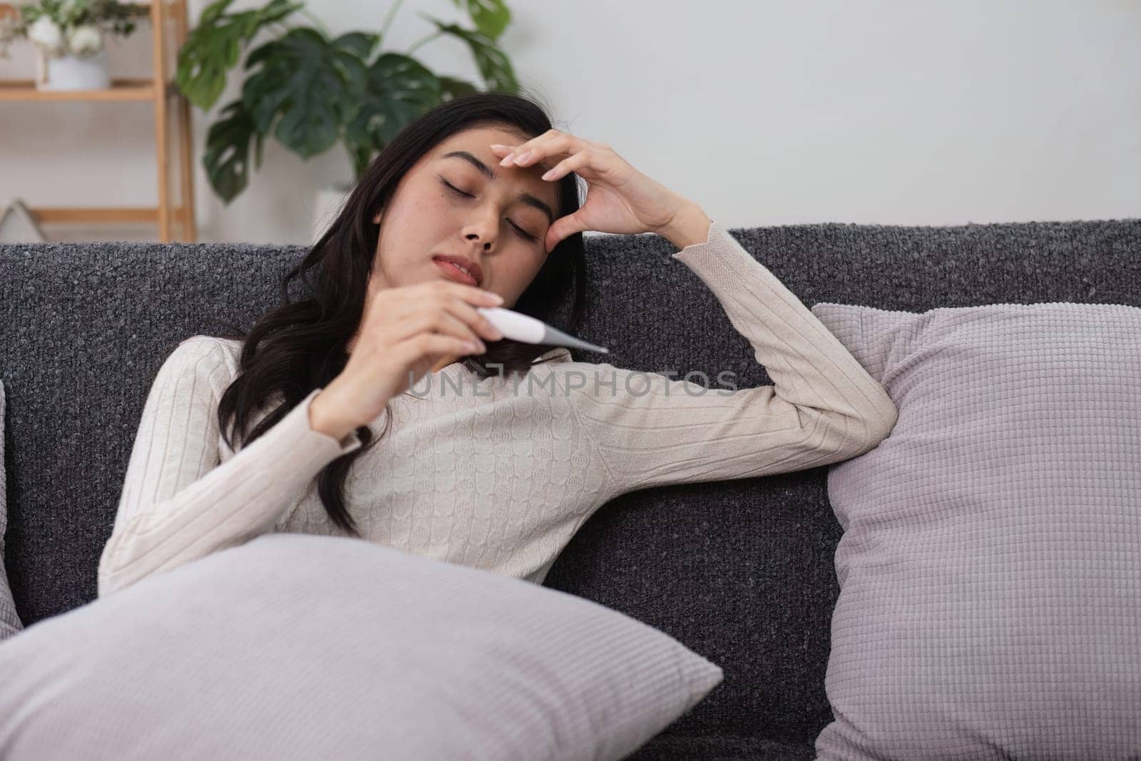 A young woman feeling unwell rests on the couch thermometer in hand embodying the struggle of sickness at home.