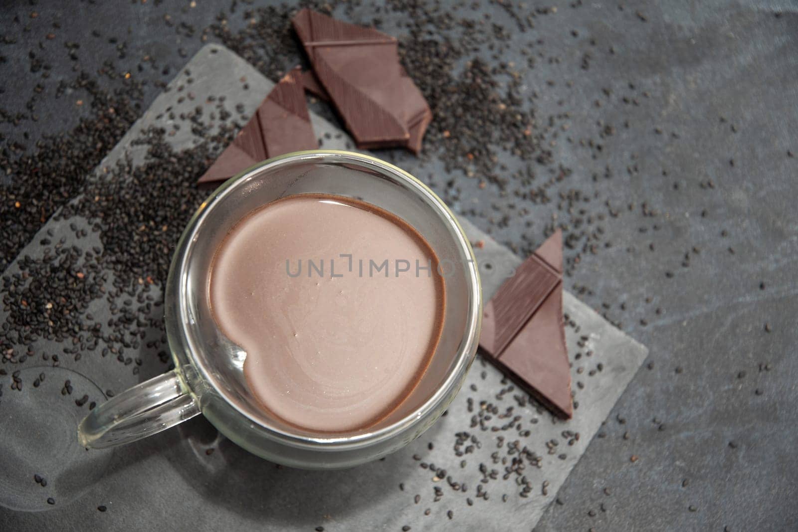 Glass cup of hot chocolate in heart shape on table surrounded by chocolate pieces and black chia seeds on grey textured background, Desert concept in color of the year 2025, mocha mousse,