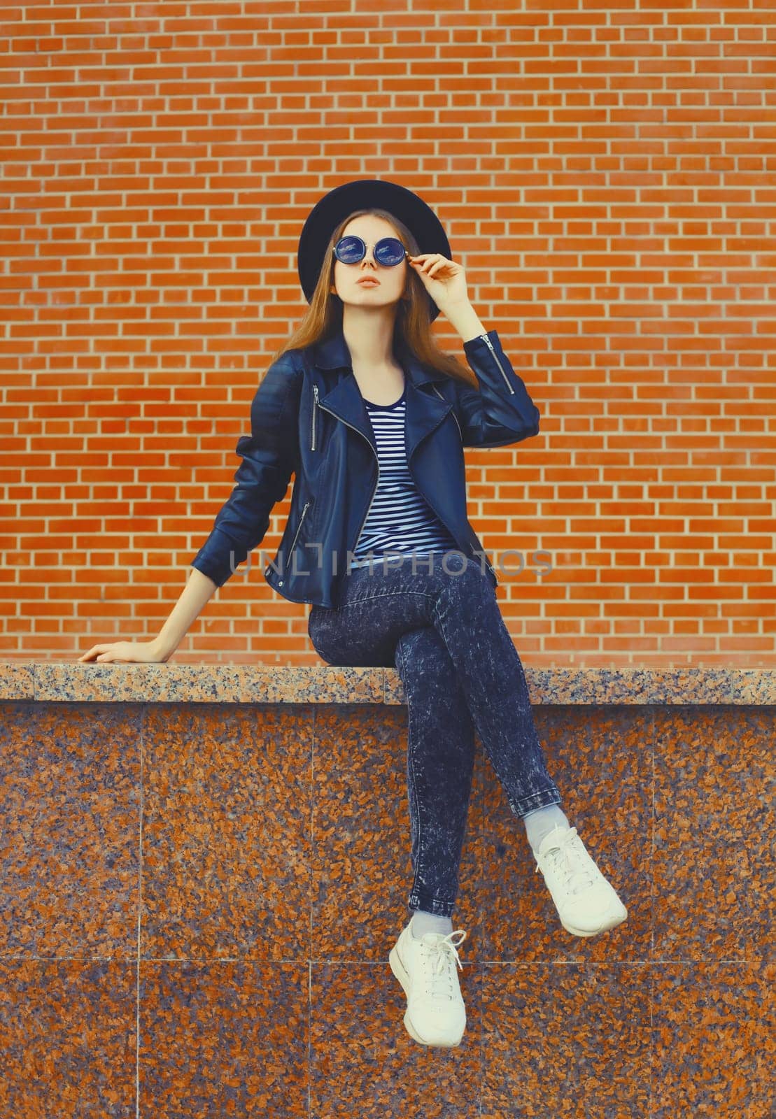 Stylish young woman model wearing black round hat, leather jacket in rock style on city street over brick wall background
