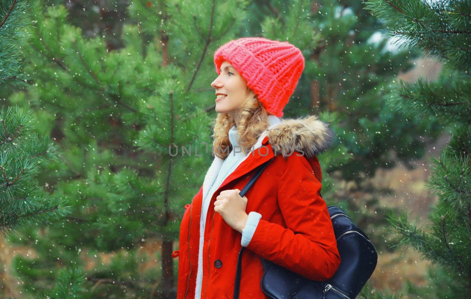 Happy young woman looking away with backpack, enjoys snow, warm weather against Christmas tree background, joyful girl in red hat standing in snowy forest with snowflakes