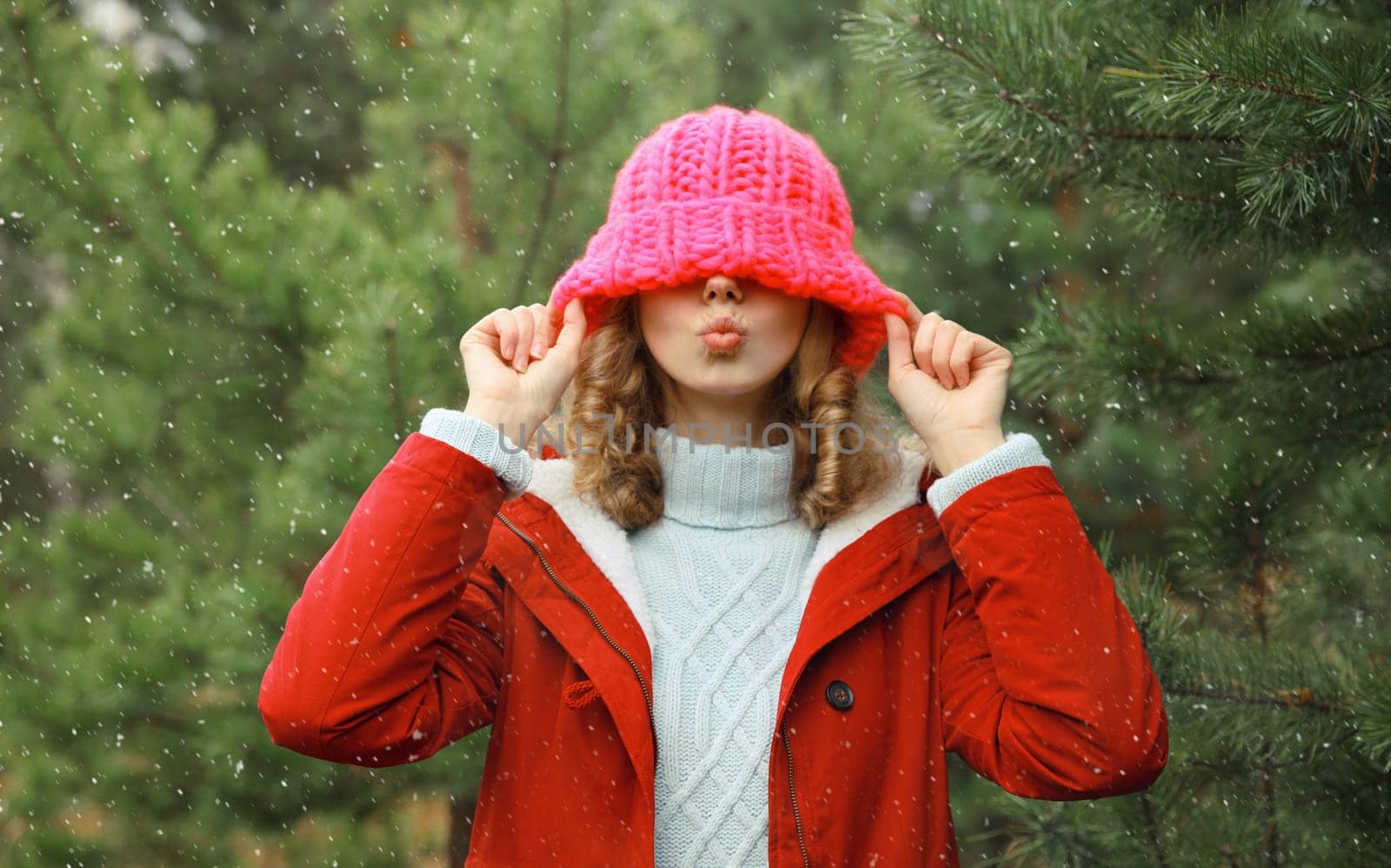 Happy cheerful young woman having fun enjoys snow, warm weather in winter park against Christmas tree, joyful girl in hat standing in forest with snowflakes, festive background
