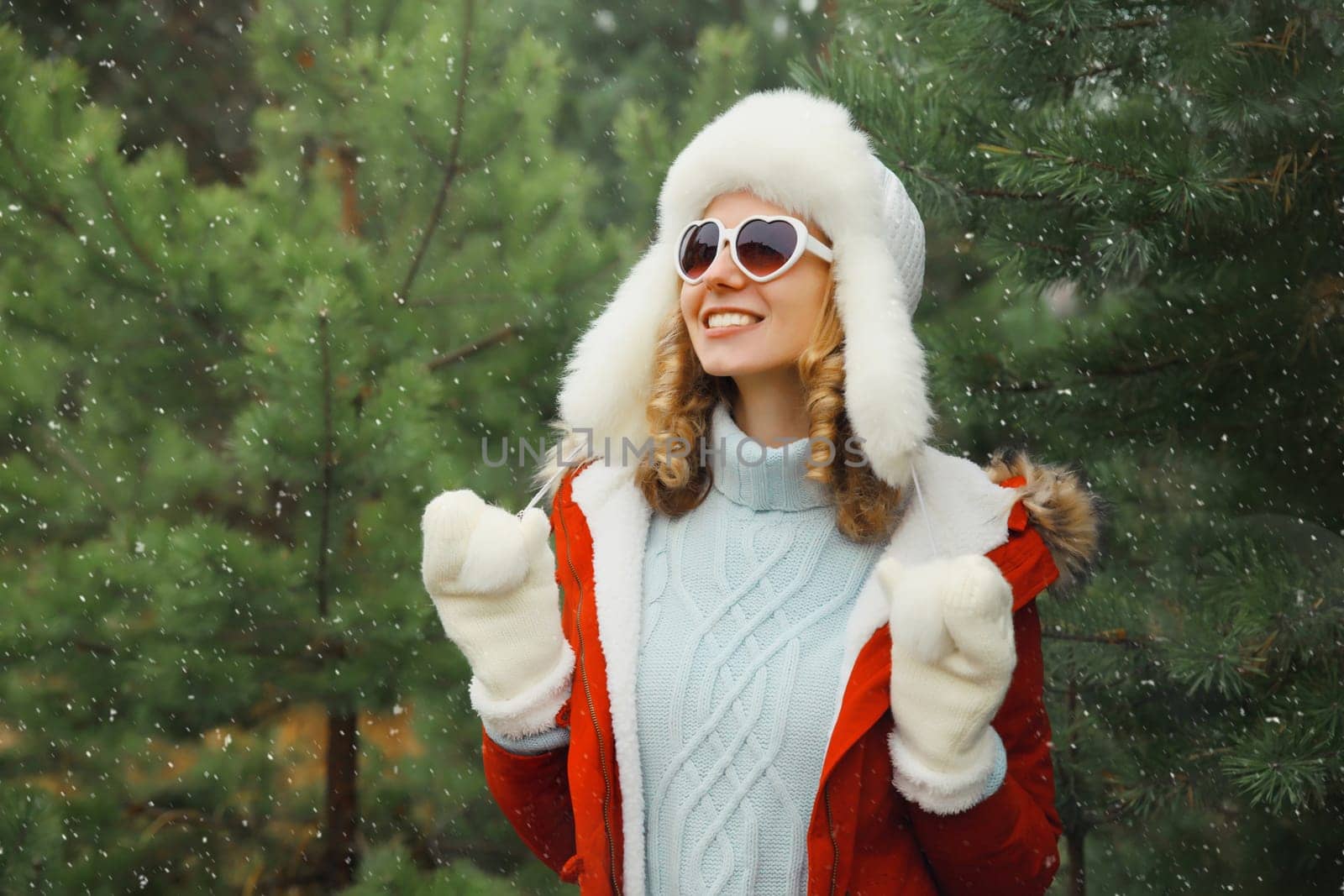 Happy smiling young woman having fun enjoys snow, warm weather in winter park against Christmas tree, joyful girl in hat, glasses standing in forest with snowflakes, festive background