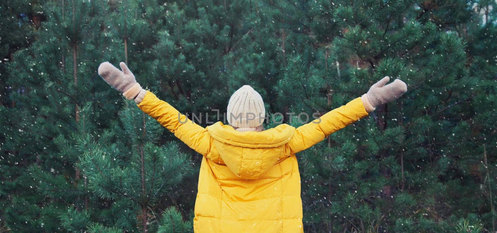 Back view, winter day, happy young woman enjoys snow, warm weather against Christmas tree, joyful girl in hat, yellow jacket raising her hands up in snowy green forest with snowflakes
