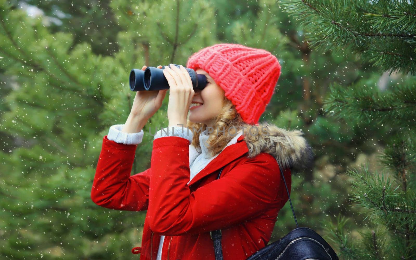 Happy woman tourist looking through binoculars watching birds, wild animals in winter forest, young female hiker enjoying nature, while walking in park