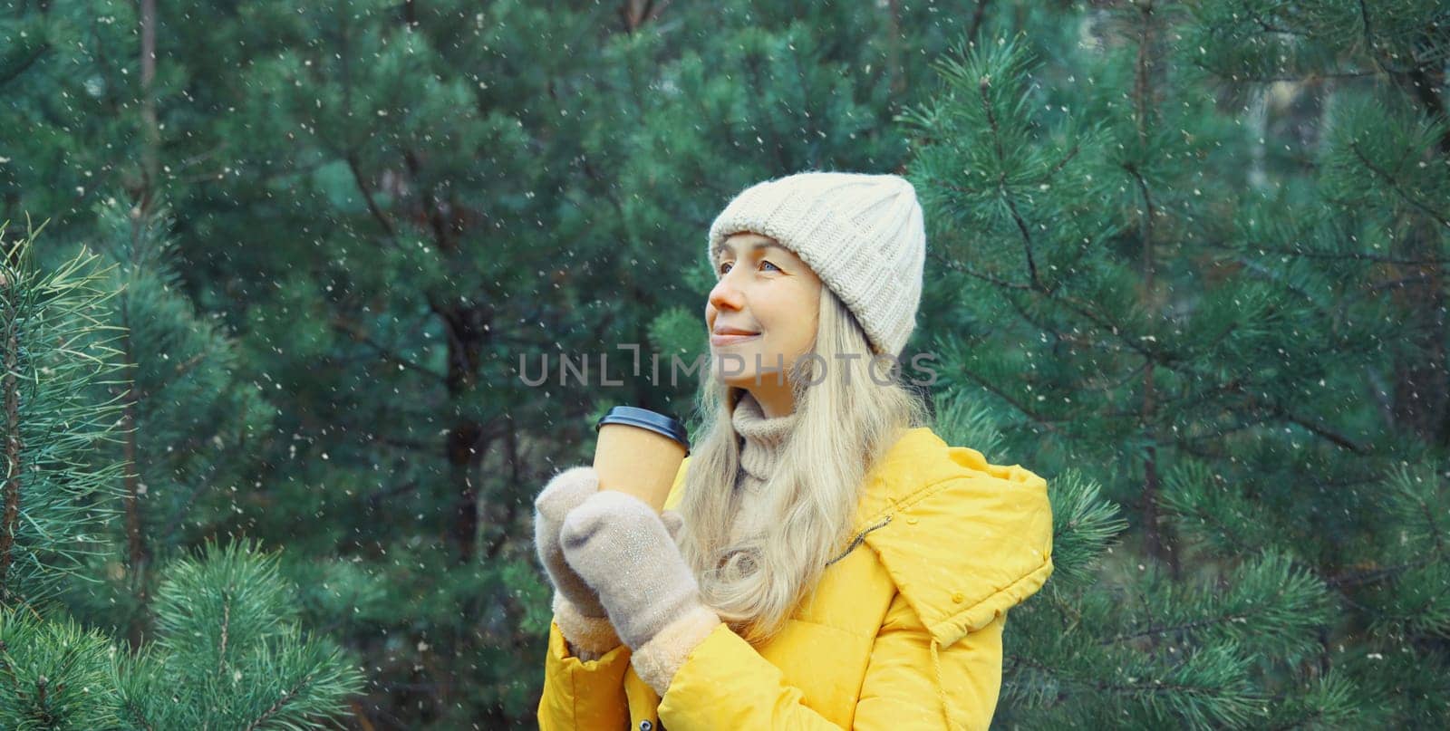 Winter day, happy smiling mature woman holding cup of coffee drink enjoys snow, warm weather against Christmas tree, wearing yellow jacket, hat, standing in snowy forest with snowflakes