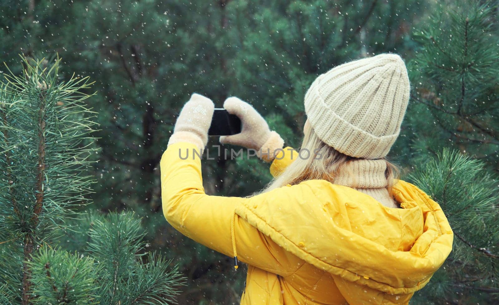 Young woman taking picture on phone Christmas tree in winter park, girl with smartphone outdoors, wearing yellow jacket, hat, standing in snowy forest with snowflakes, nature background