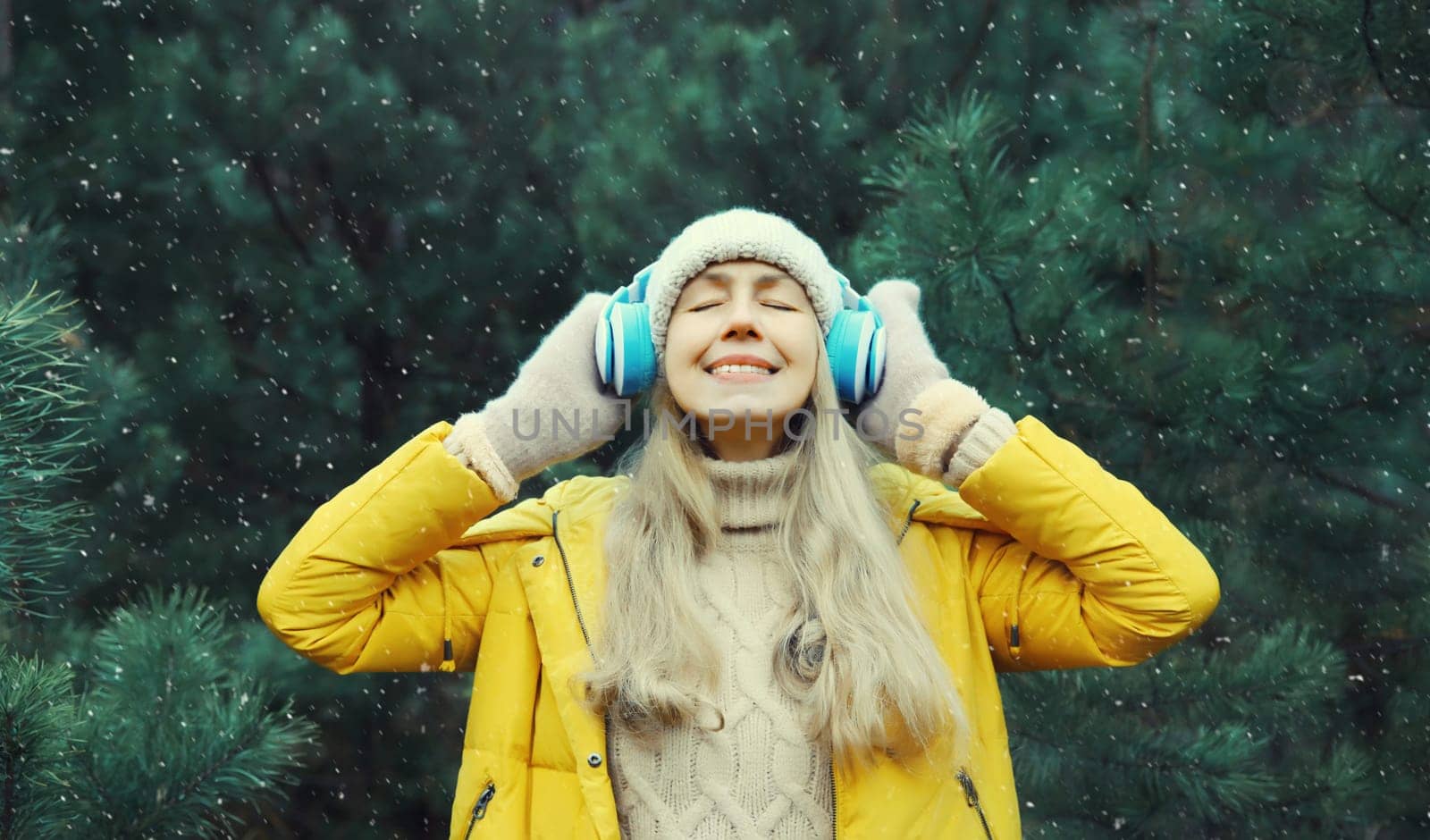 Portrait of happy modern young woman enjoying listening to music with headphones in hat, bright yellow jacket in winter forest against Christmas tree background with snow
