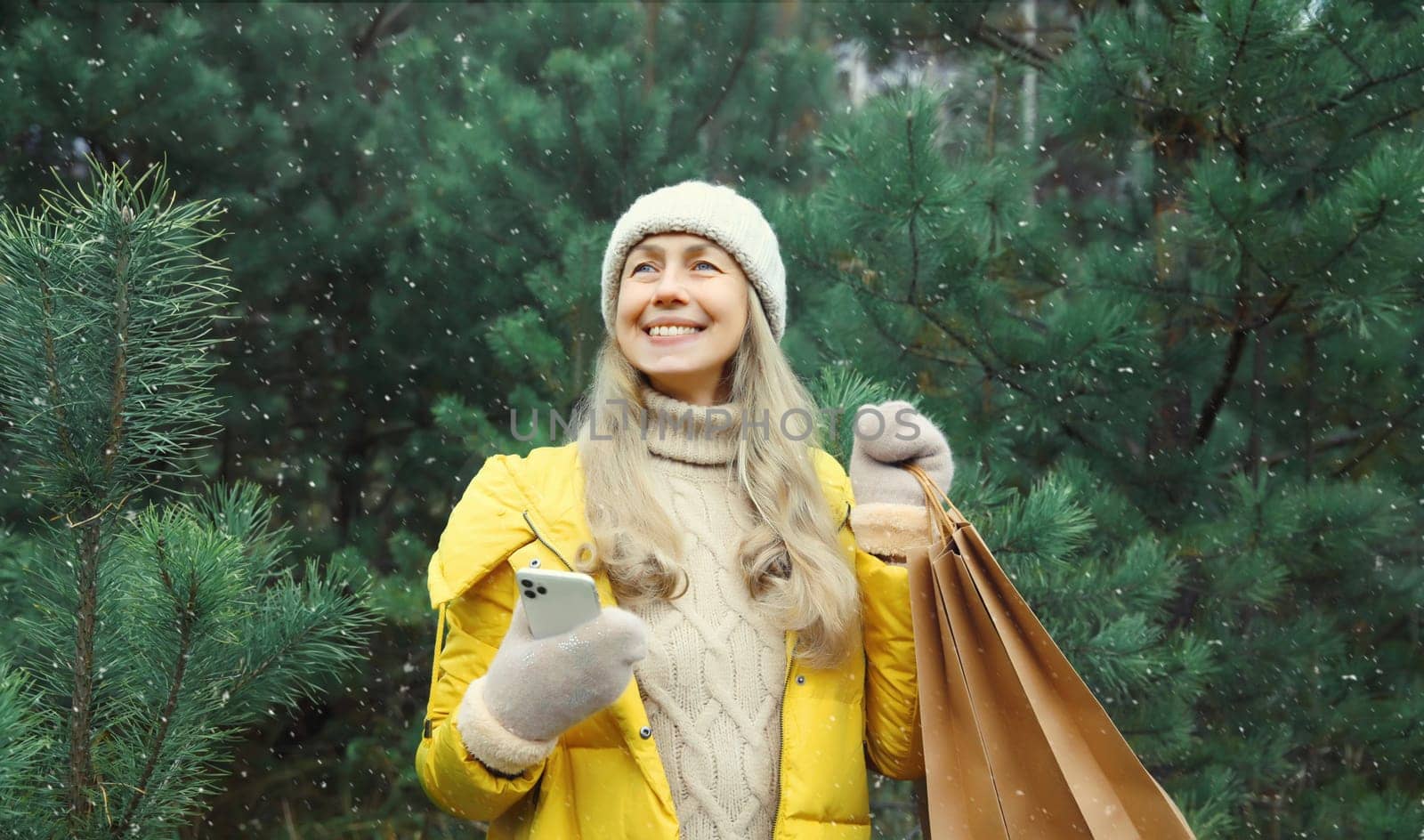 Happy smiling mature woman with phone, shopping bag in winter against Christmas tree, joyful girl using smartphone in white hat, yellow jacket standing in snowy forest