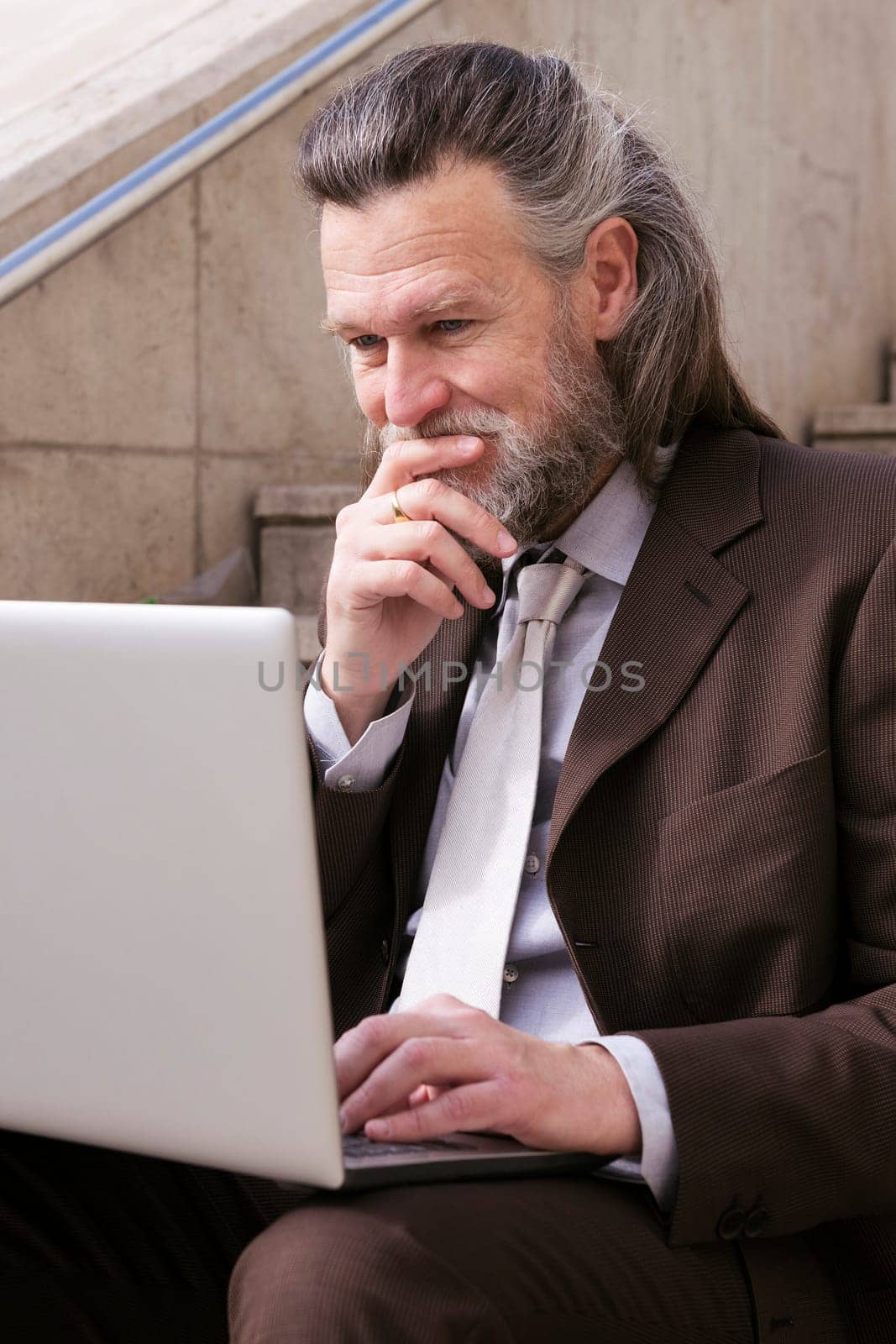 pensive mature male entrepreneur in elegant suit siting on urban stairway and working online on laptop in city, concept of business and middle age man lifestyle