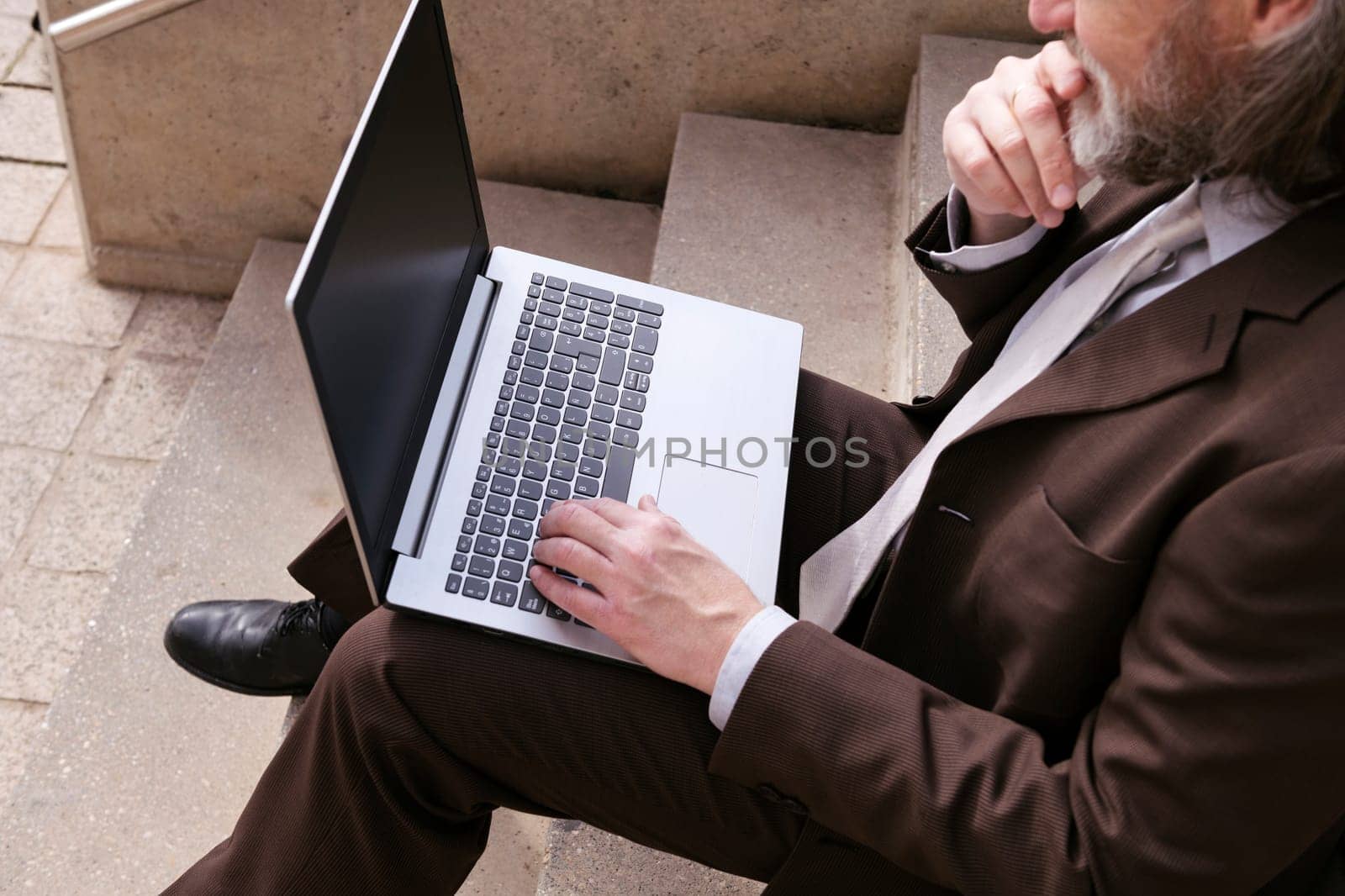 crop of unrecognizable male entrepreneur in formal clothes sitting on urban stairway and working online on laptop in city, concept of business and remote work
