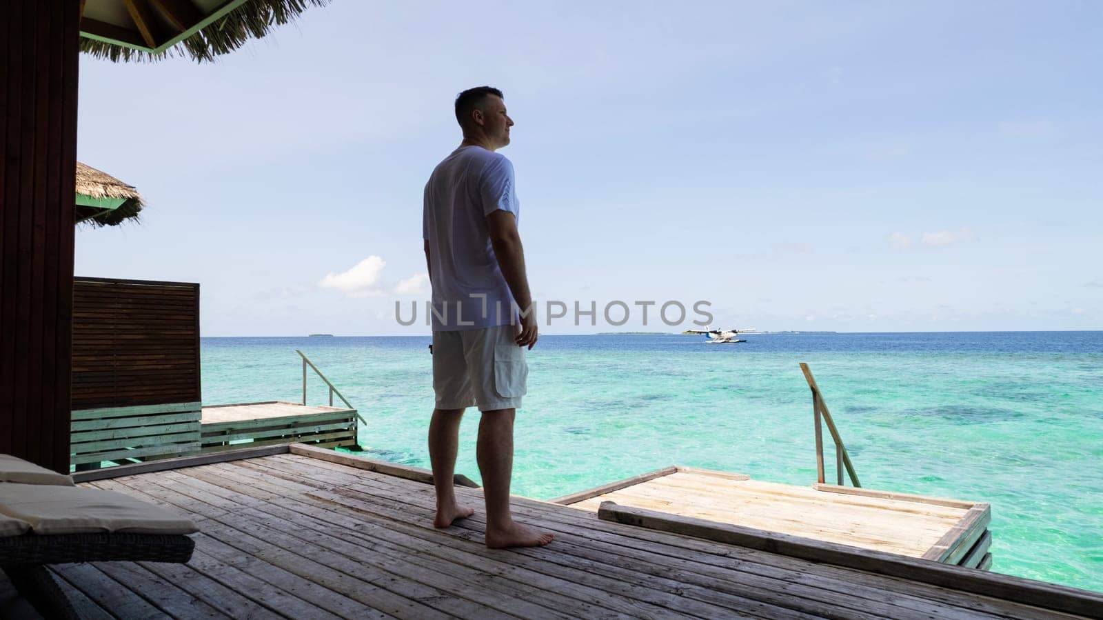 A guy in white clothes admires the turquoise ocean. Vacation in the Maldives. He looks into the future and enjoys it. Small waves. Descent into the water. Wooden villa. Bungalows on the water