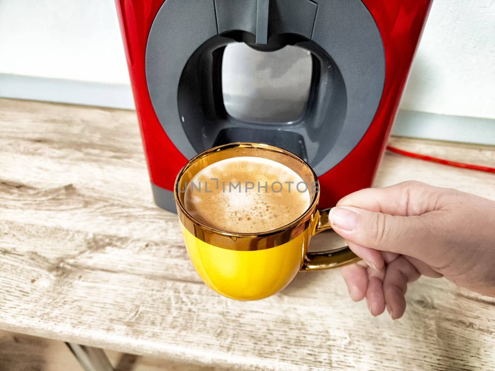 A person holds a golden coffee mug filled with freshly brewed coffee near a sleek home coffee machine on a wooden surface.