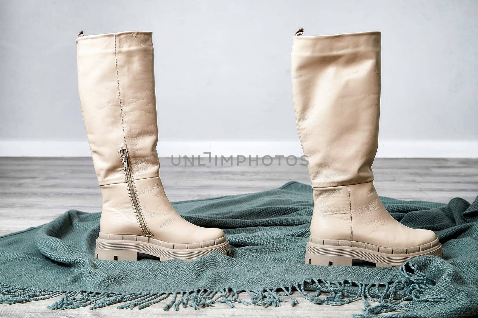 Model poses with fashionable knee-high boots on a textured green blanket in a bright, minimalist interior space.