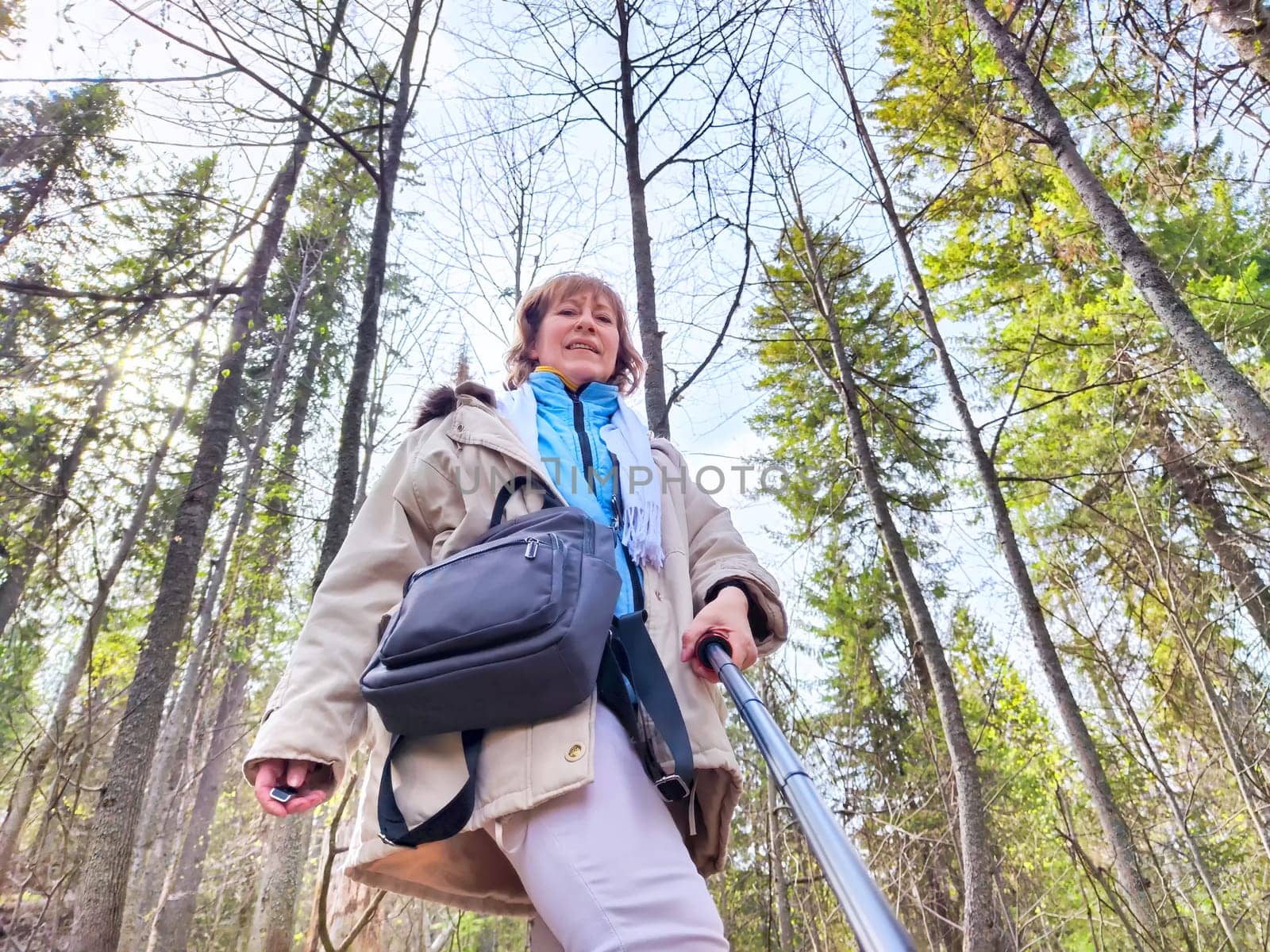 A woman explores a lush forest, capturing the beauty of nature with her selfie stick on a bright day. The adventures of a middle-aged female tourist on a trip
