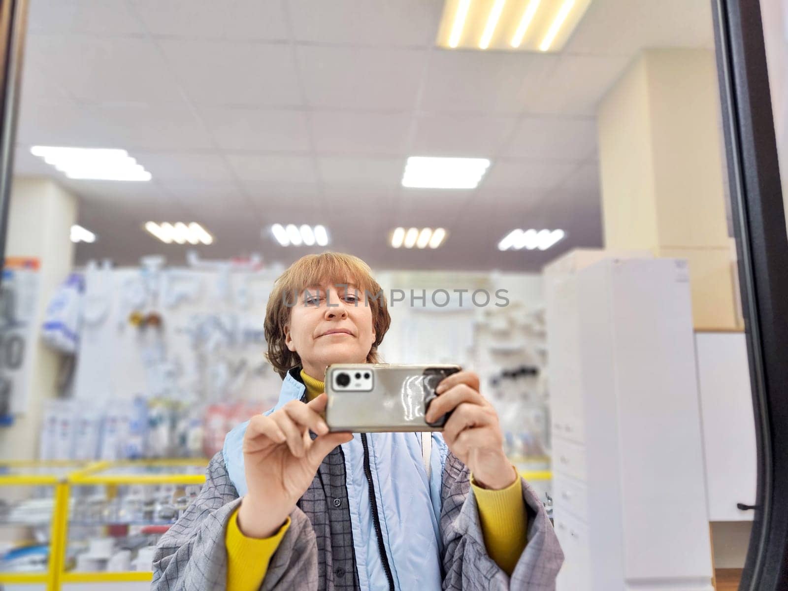 A middle-aged woman captures a selfie in a well-lit store with shelves full of household goods, smiling confidently at the camera.