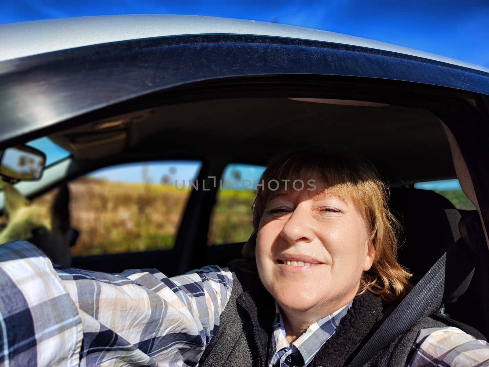 A middle-aged woman smiles happily as she takes a selfie in her car while driving through a picturesque countryside landscape.
