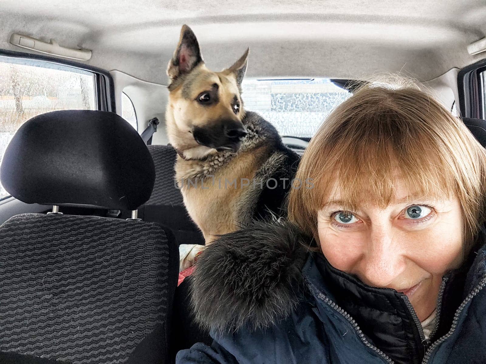 A middle-aged woman smiles for a selfie while her dog sits in the backseat, capturing their adventure spirit together.