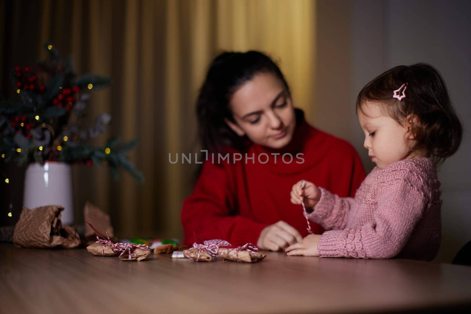 mother and her little child girl together wrapping Christmas gifts at home in the evening, preparing presents for family.