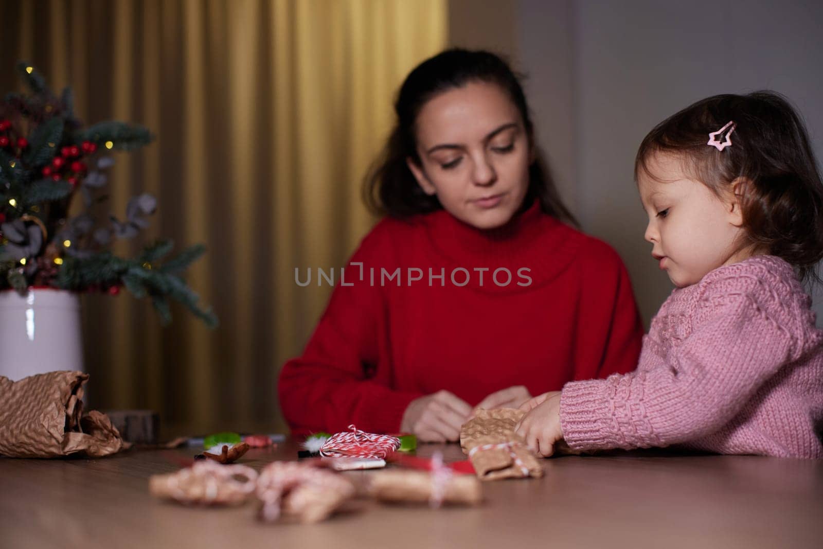 mother and her little child girl together wrapping Christmas gifts at home in the evening, preparing presents for family.