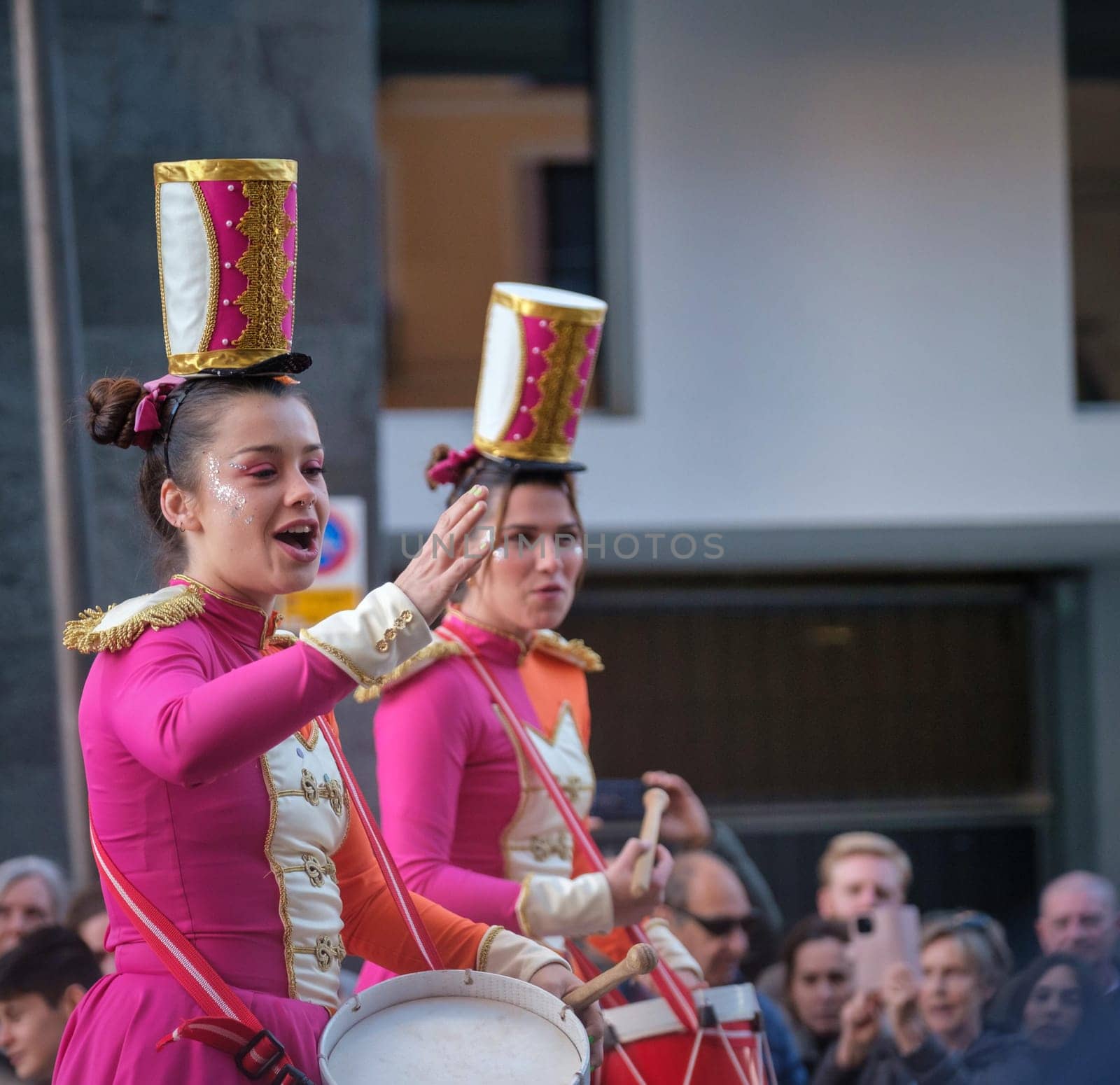 Performers in pink military-style costumes during an outdoor event. One salutes while the other plays a snare drum, thrilling the spectator-filled street