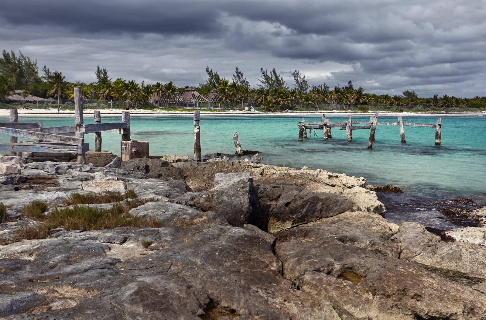 Decaying wooden pier stretches into the turquoise waters of the caribbean sea, under a dramatic cloudy sky in xpu-ha, mexico