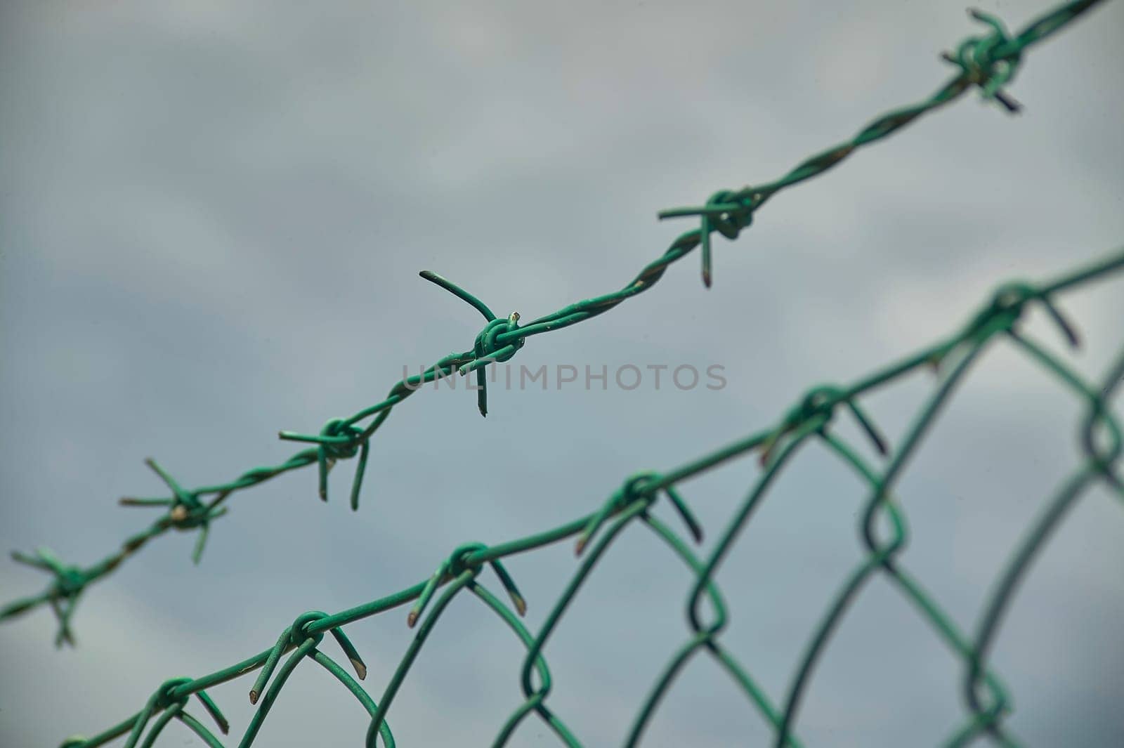 Close-up of green barbed wire atop a chain link fence, suggesting security and restricted access against a cloudy sky