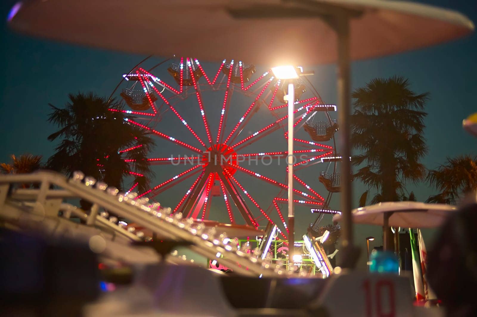 Red illuminated ferris wheel rotating at night in amusement park with palm trees and other attractions