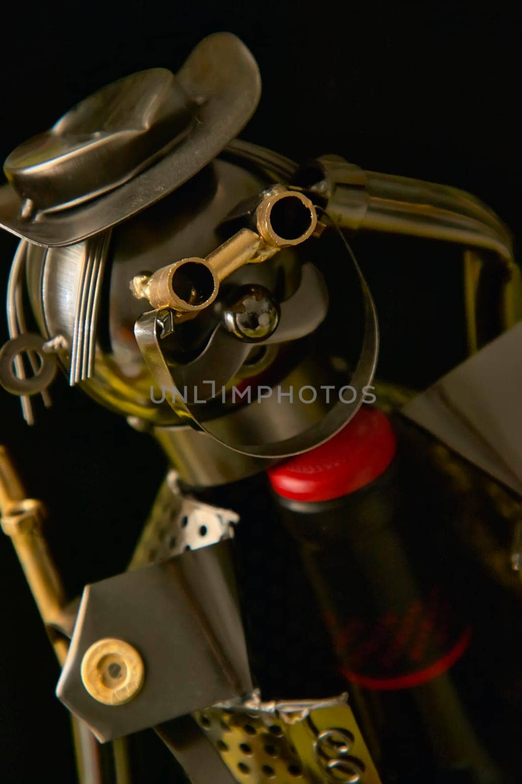 Metal sculpture of a figure wearing a hat and goggles, holding a wine bottle, on a black background