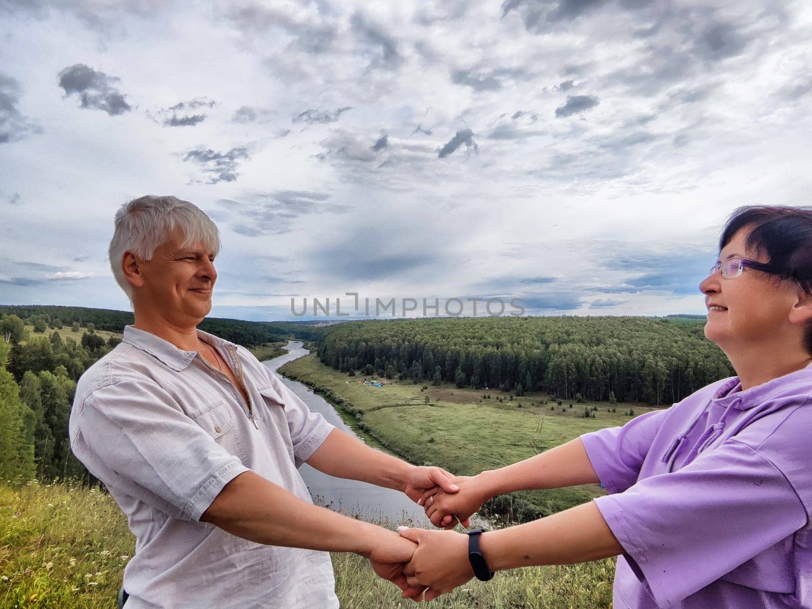 A couple shares a happy moment, holding hands and smiling as they overlook a peaceful river valley under a cloudy sky.