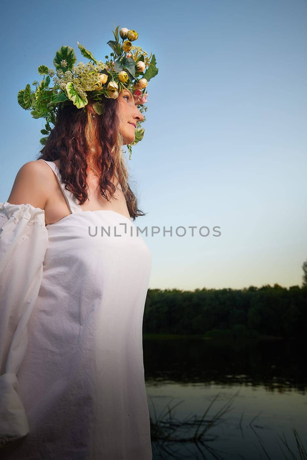 Slavic plump chubby girl in long white dress on the feast of Ivan Kupala with flowers and water in a river or lake on summer evening